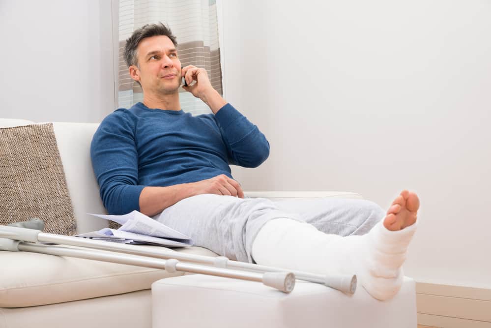 Accident insurance - Man with broken leg sitting on the couch talking on the phone.
